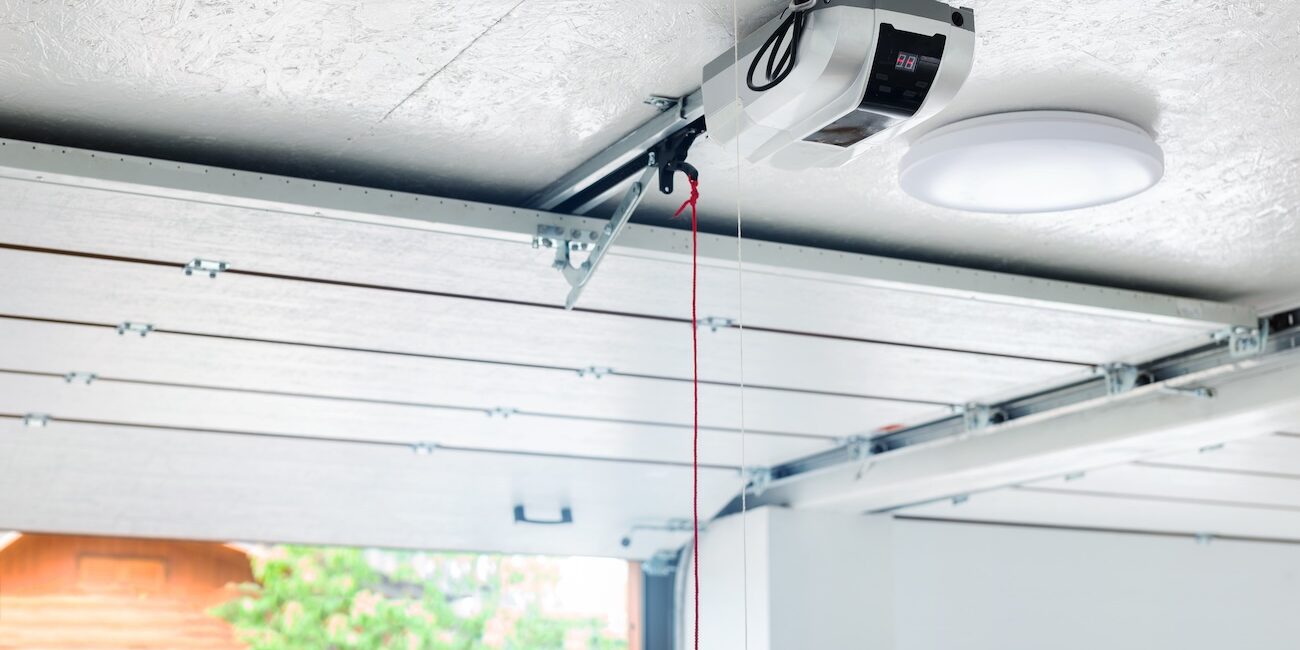 Opening door and automatic garage door opener electric engine gear mounted on ceiling with emergency cord. Double place empty garage interior with rolling entrance gate Opening door and automatic garage door opener electric engine gear mounted on ceiling with emergency cord. Double place empty garage interior with rolling entrance gate.
