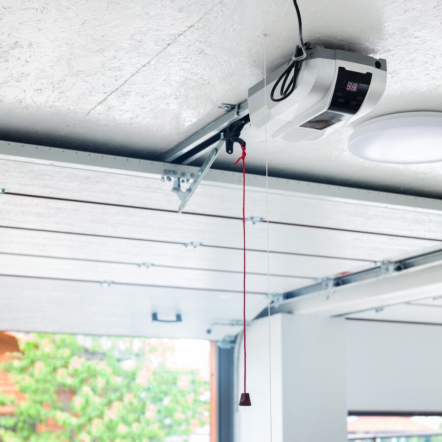 Opening door and automatic garage door opener electric engine gear mounted on ceiling with emergency cord. Double place empty garage interior with rolling entrance gate Opening door and automatic garage door opener electric engine gear mounted on ceiling with emergency cord. Double place empty garage interior with rolling entrance gate.